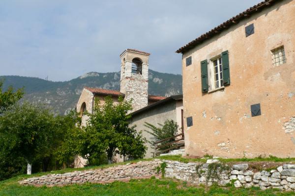 esterno edificio antico ocra con scuri verdi, campanile sullo sfondo, cortile con piante e muro di supporto in pietra