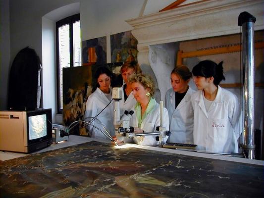 studendi e insegnante con camici bianchi attorno tavolo con dipinto e macchinari per il restauro, antico camino in muratura sullo sfondo