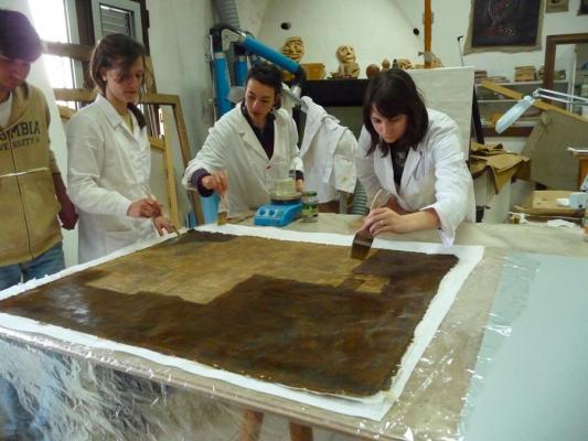 studendi con camici bianchi dentro laboratorio, intenti a restaurare un dipinto posto su un tavolo con pennelli, camino in muratura sullo sfondo, statue umanoidi sul camino