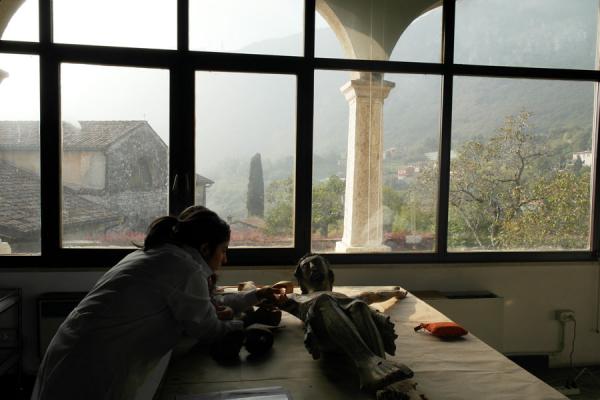 studente con camice intento a restaurare statua di Cristo crocefisso, finestre a tutta parete, sfondo antico edificio in sasso, colonna e vallata con alberi