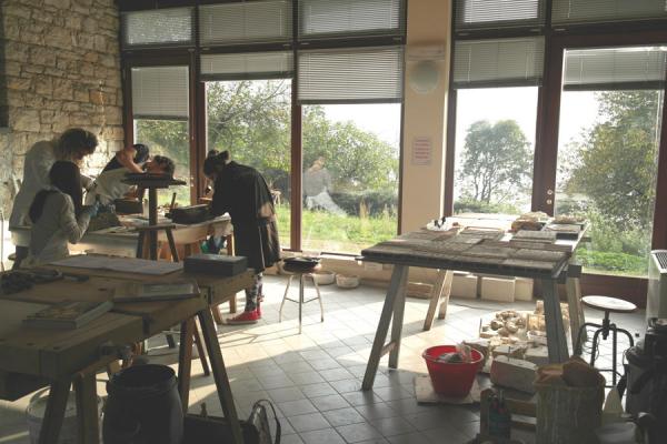 studenti con camici intenti a lavorare su pietre, laboratorio con tre tavoli, pietre e strumenti per il restauro, finestre a parete su un lato della stanza, che si affacciano su cortile con prato e alberi